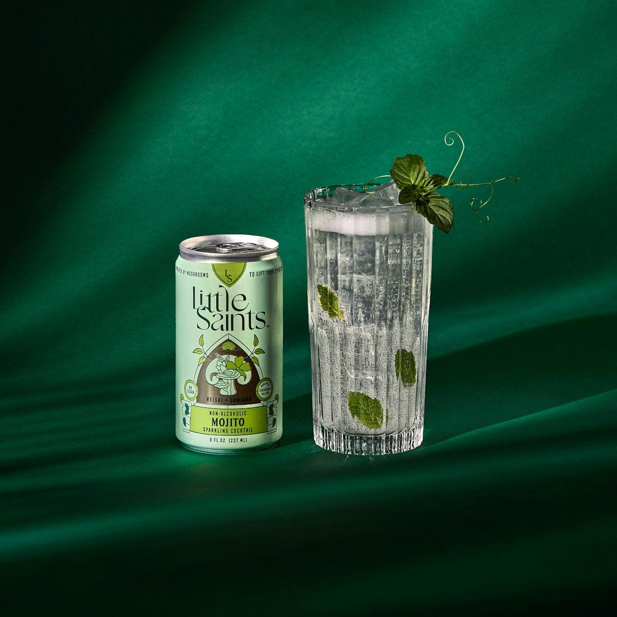 A can of Little Snaps Mint Lemon sparkling drink stands next to a clear textured glass filled with ice and mint leaves, set against a dark green draped fabric background with dramatic lighting.