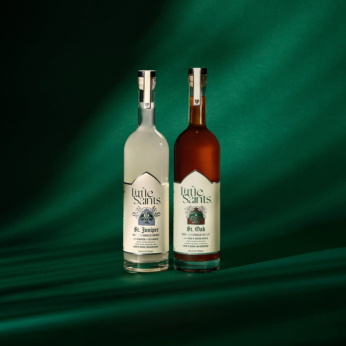 Two bottles of Little Snips spirits, one clear and one amber-brown, standing side by side on a dark green draped fabric background with dramatic lighting.